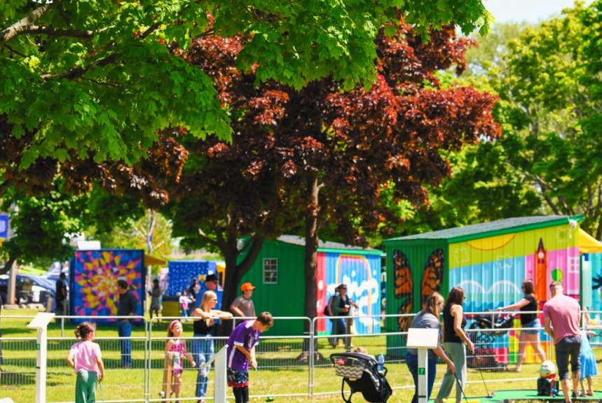 Families enjoying a mini-golf game at Pop-Ups on the Bay in West Zwick’s Park, Belleville. The vibrant scene includes children and adults playing under lush green trees, with colorful murals in the background. A stroller is also visible, emphasizing the family-friendly atmosphere.